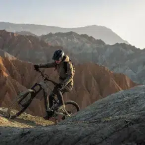 A helmeted cyclist rides a mountain bike along a rocky ridge, with layered, barren mountains stretching across the background.
