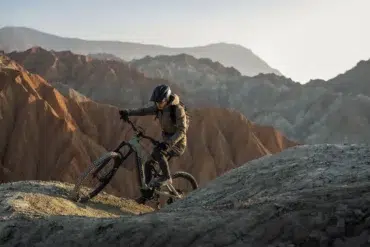A helmeted cyclist rides a mountain bike along a rocky ridge, with layered, barren mountains stretching across the background.