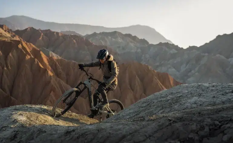A helmeted cyclist rides a mountain bike along a rocky ridge, with layered, barren mountains stretching across the background.