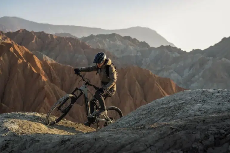 A helmeted cyclist rides a mountain bike along a rocky ridge, with layered, barren mountains stretching across the background.