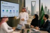 Business meeting with four people in traditional Saudi attire and a suited man, beside a screen displaying Microsoft Dynamics 365.