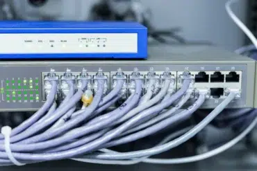 Network equipment with numerous Ethernet cables plugged into a 24-port switch beneath a blue wireless router.