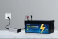 A 12.8-volt 210Ah LiFePO4 battery connected to a silver inverter and wall outlet, with red and black clamps attached.