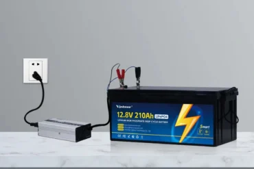 A 12.8-volt 210Ah LiFePO4 battery connected to a silver inverter and wall outlet, with red and black clamps attached.
