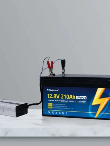 A 12.8-volt 210Ah LiFePO4 battery connected to a silver inverter and wall outlet, with red and black clamps attached.