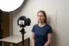 Woman in a navy T-shirt sits against a plain backdrop while a camera on a tripod and softbox light set up a portrait shoot.