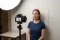 Woman in a navy T-shirt sits against a plain backdrop while a camera on a tripod and softbox light set up a portrait shoot.