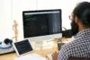 A bearded man in glasses works at a desk with code displayed on a desktop monitor and tablet.