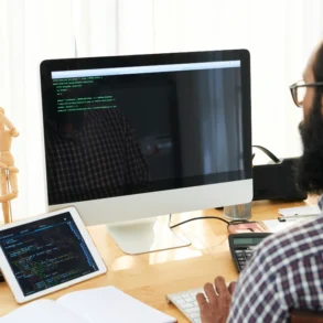 A bearded man in glasses works at a desk with code displayed on a desktop monitor and tablet.