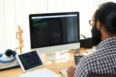 A bearded man in glasses works at a desk with code displayed on a desktop monitor and tablet.