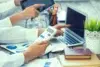 Two people work at a desk with a laptop, tablet, calculator, charts, notebooks, pens, and a potted plant.