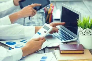 Two people work at a desk with a laptop, tablet, calculator, charts, notebooks, pens, and a potted plant.