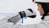 A helmeted skier in white gear checks a wristwatch on a snowy slope, with a blue safety net behind him; the scene feels focused and calm.