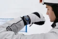 A helmeted skier in white gear checks a wristwatch on a snowy slope, with a blue safety net behind him; the scene feels focused and calm.