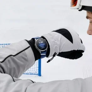 A helmeted skier in white gear checks a wristwatch on a snowy slope, with a blue safety net behind him; the scene feels focused and calm.