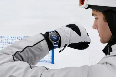 A helmeted skier in white gear checks a wristwatch on a snowy slope, with a blue safety net behind him; the scene feels focused and calm.