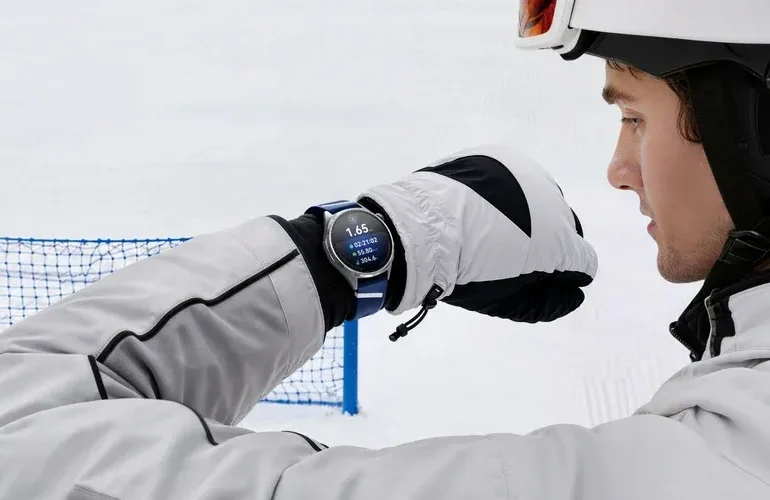 A helmeted skier in white gear checks a wristwatch on a snowy slope, with a blue safety net behind him; the scene feels focused and calm.