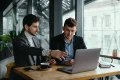 Two men in business attire discuss a laptop at a café table by large windows, with coffee and a smartphone nearby; collaborative, focused mood.