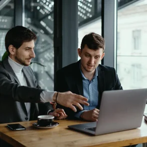 Two men in business attire discuss a laptop at a café table by large windows, with coffee and a smartphone nearby; collaborative, focused mood.