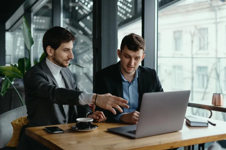 Two men in business attire discuss a laptop at a café table by large windows, with coffee and a smartphone nearby; collaborative, focused mood.