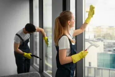 Two cleaners in gloves and dark overalls clean large windows, with one wiping glass using a squeegee and the other vacuuming frames.