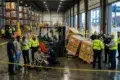 Industrial warehouse safety alert: investigators gather around an injured worker and forklift near fallen crates, behind yellow caution tape at Chesapeake port.