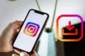 Hand holding a smartphone displaying the Instagram logo beside a colorful download icon, suggesting saving social media content; bright, modern tone.