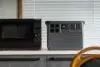 Black microwave beside a portable power station on a kitchen counter, with window blinds behind and a wooden chair in the foreground.