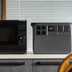 Black microwave beside a portable power station on a kitchen counter, with window blinds behind and a wooden chair in the foreground.