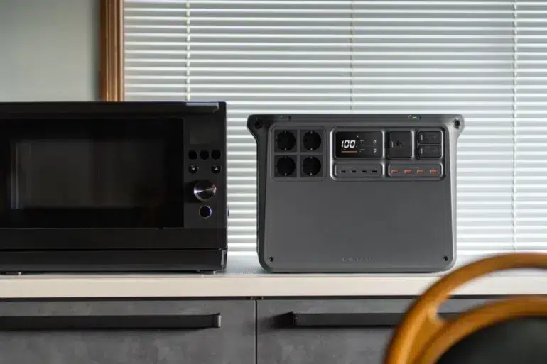 Black microwave beside a portable power station on a kitchen counter, with window blinds behind and a wooden chair in the foreground.