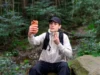 A man in a black cap and outdoor clothing sits on a rock in the forest, holding up an orange smartphone and adjusting his neck gaiter.