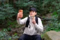 A man in a black cap and outdoor clothing sits on a rock in the forest, holding up an orange smartphone and adjusting his neck gaiter.