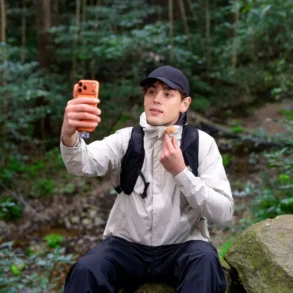 A man in a black cap and outdoor clothing sits on a rock in the forest, holding up an orange smartphone and adjusting his neck gaiter.