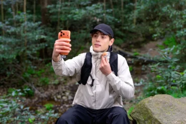 A man in a black cap and outdoor clothing sits on a rock in the forest, holding up an orange smartphone and adjusting his neck gaiter.