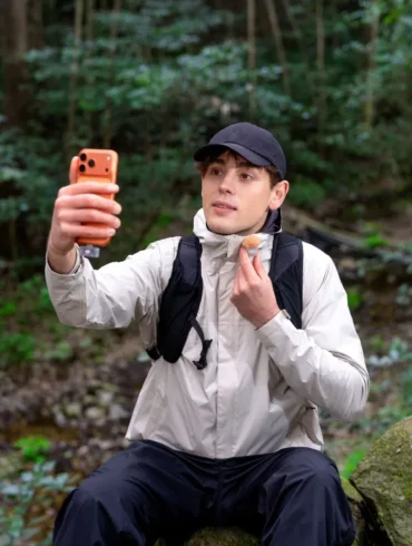 A man in a black cap and outdoor clothing sits on a rock in the forest, holding up an orange smartphone and adjusting his neck gaiter.