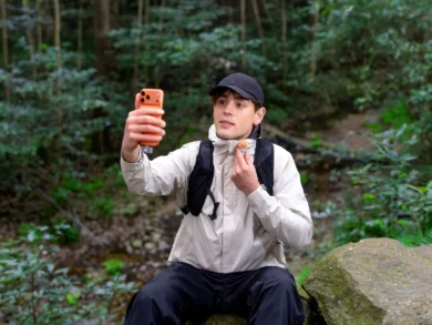A man in a black cap and outdoor clothing sits on a rock in the forest, holding up an orange smartphone and adjusting his neck gaiter.