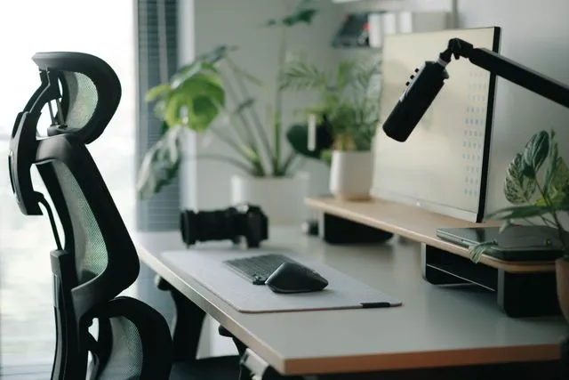Modern minimalist office with an ergonomic black chair, wide desk, monitor, adjustable lamp, keyboard, and plants, creating a clean, calm workspace.