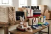 Stacks of reusable canvas tote bags in neutral and bright colors cover wooden tables in a sunlit warehouse, creating a busy, well-stocked feel.