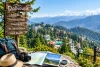 A wooden Dalhousie sign lists nearby sights above a map, guidebook, compass, hat, and backpack, overlooking a sunny mountain town and snow-capped Himalayas.