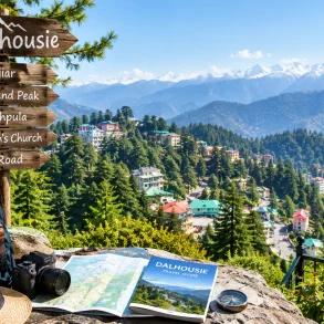 A wooden Dalhousie sign lists nearby sights above a map, guidebook, compass, hat, and backpack, overlooking a sunny mountain town and snow-capped Himalayas.