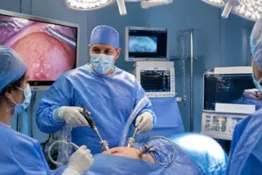 Surgeon and masked medical team perform minimally invasive surgery in a brightly lit operating room, with monitors showing the procedure; focused, clinical atmosphere.