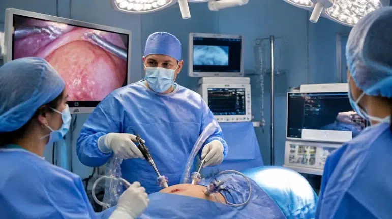 Surgeon and masked medical team perform minimally invasive surgery in a brightly lit operating room, with monitors showing the procedure; focused, clinical atmosphere.
