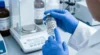 In a bright laboratory, blue-gloved hands hold and point to a labeled chemical bottle beside a digital scale, conveying careful, precise work.