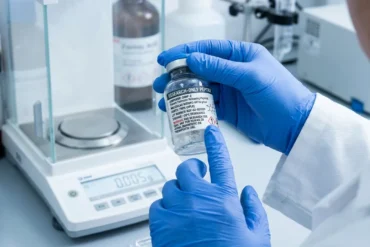 In a bright laboratory, blue-gloved hands hold and point to a labeled chemical bottle beside a digital scale, conveying careful, precise work.