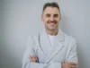 A smiling Serhii Tokarev in a white coat stands with arms crossed against a plain gray background, conveying confidence and warmth.