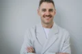 A smiling Serhii Tokarev in a white coat stands with arms crossed against a plain gray background, conveying confidence and warmth.