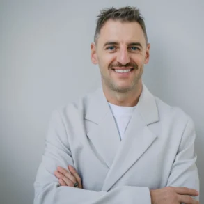 A smiling Serhii Tokarev in a white coat stands with arms crossed against a plain gray background, conveying confidence and warmth.