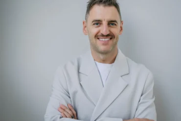 A smiling Serhii Tokarev in a white coat stands with arms crossed against a plain gray background, conveying confidence and warmth.