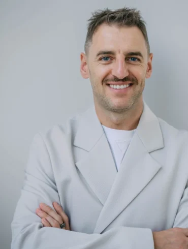 A smiling Serhii Tokarev in a white coat stands with arms crossed against a plain gray background, conveying confidence and warmth.