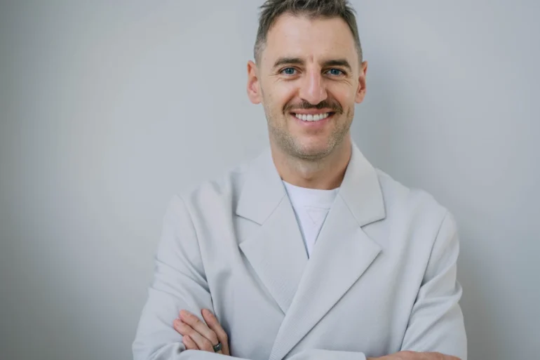 A smiling Serhii Tokarev in a white coat stands with arms crossed against a plain gray background, conveying confidence and warmth.